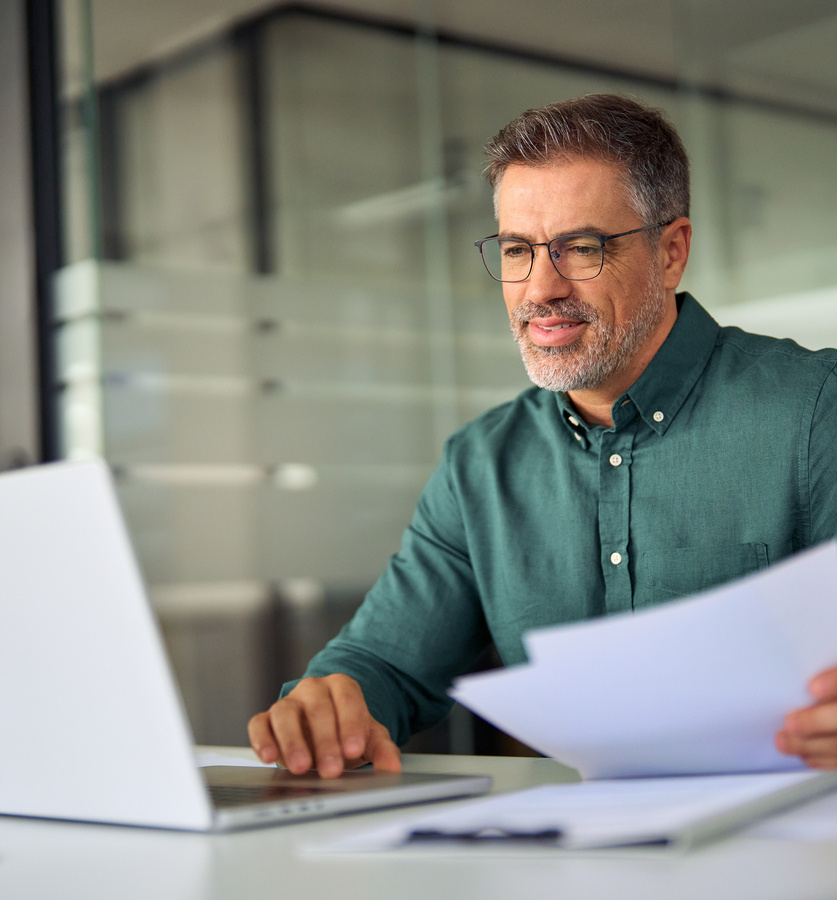 Smiling middle aged executive holding documents using laptop in office at desk. Prozesse digitalisieren