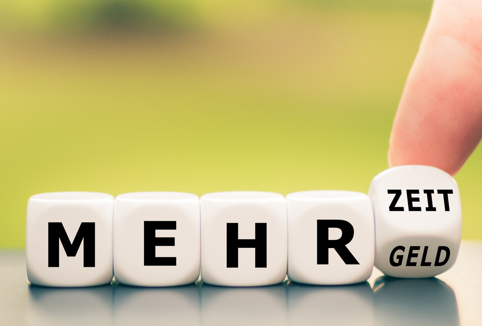 Hand turns dice and changes the German expression "mehr Geld" ("more money") to "mehr Zeit" ("more time").